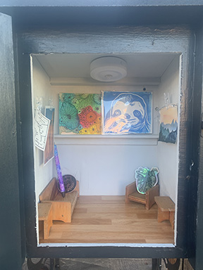 photograph of inside the free tiny art gallery featuring watercolor art, stickers, a screen printed sloth and a resin pen