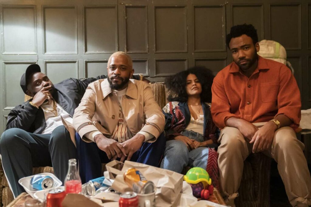 three black men and a black woman sit on a sofa with lots of food and trash on a coffee table in front of them