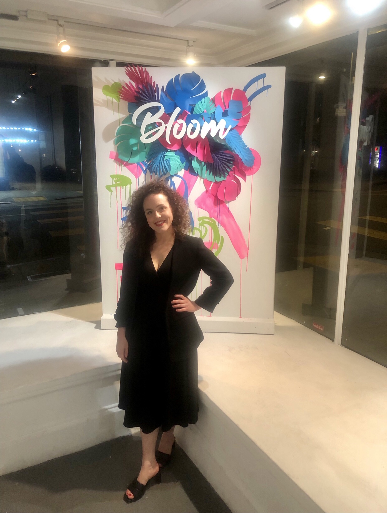 Owner and curator Ashley Voss beaming after yet another successful opening at Voss Gallery stands in front of sign that says Bloom