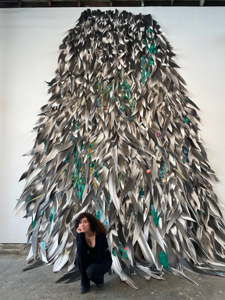 a dark haired woman sits below a large collage art piece which looks like feathers