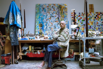 older man sits on a stool. behind him are large works of art and art supplies