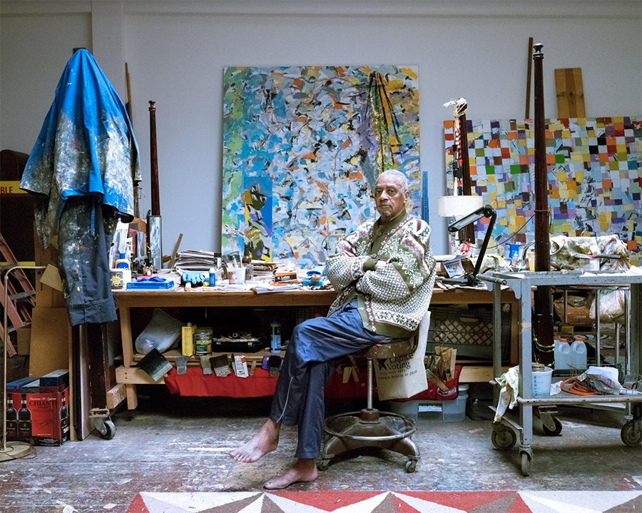 older man sits on a stool. behind him are large works of art and art supplies