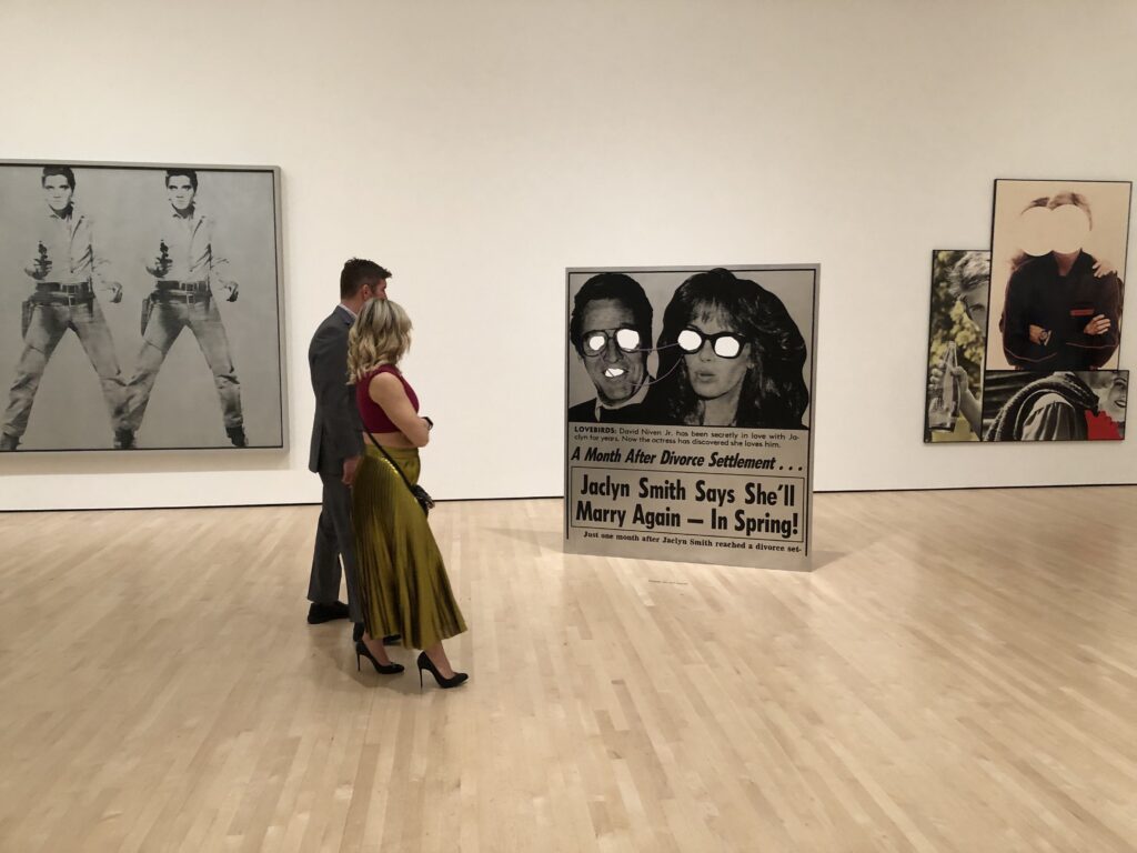 A man in a suit and a woman in a long gold skirt and high heels stroll through an exhibit of pop art 