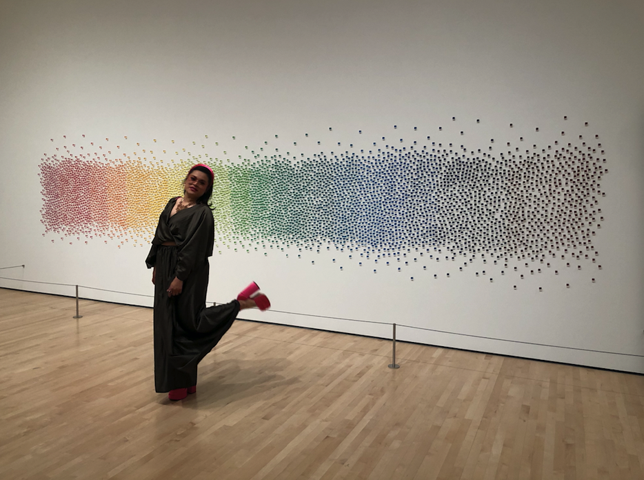 Artist Sicerra poses in an olive jumpsuit in front of a large rectangular piece of art in rainbow colors comprised of thousands of pegs stuck on the wall 