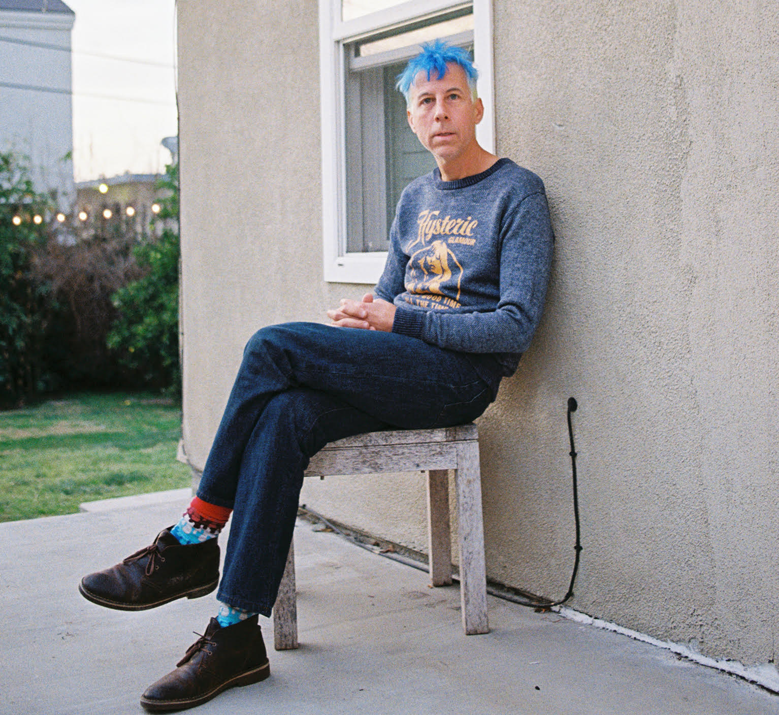 Man with blue hair sits outside on a stool