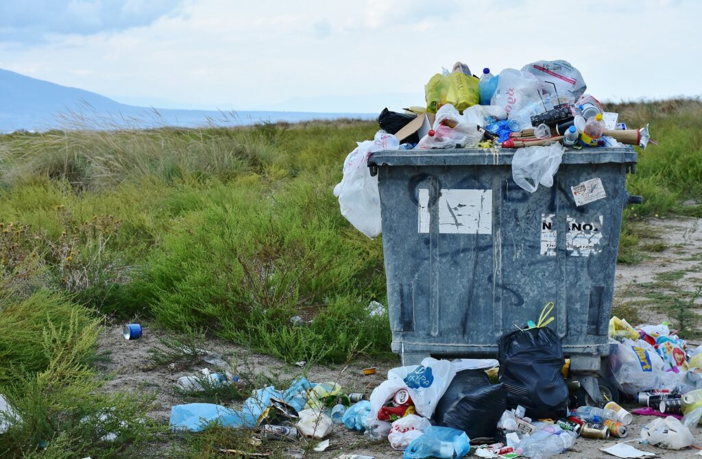 Photo of a garbage can overfilled with more garbage piled around it in an empty field.