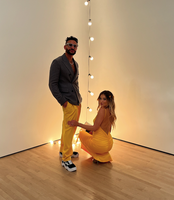Description: A couple, with the man in yellow pants and the woman in a matching yellow dress pose in front of a light sculpture. 