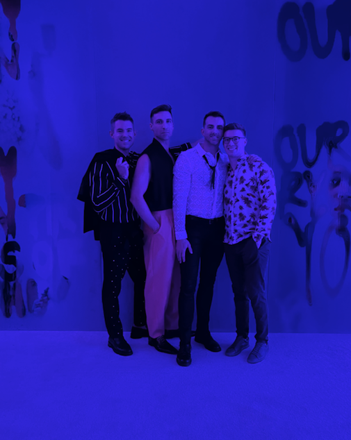 Description: Four men pose in blue lighting wearing a variety of navy, gray, pink, and white outfits.