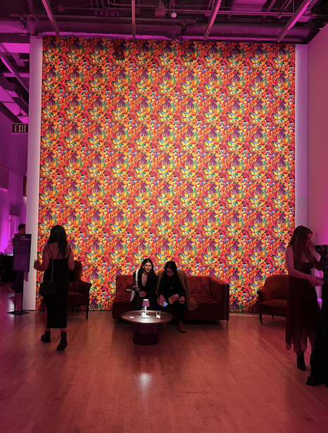 Pictured: An uplit psychedelic patterned wall with red lounge furniture and party goers mingling.