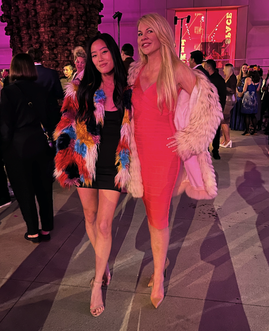 Pictured: Two women wearing short cocktail dresses pose wearing colorful oversized furry jackets.