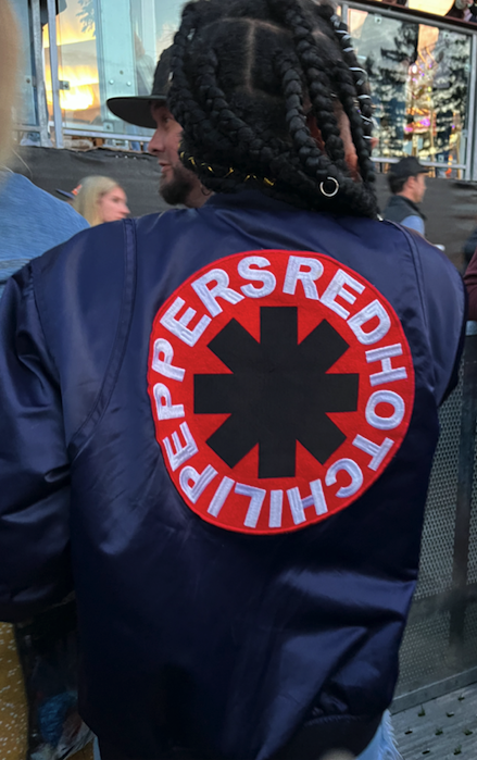 a femme leaning person with braids shows the back of her blue bomber jacket: a large red hot chili peppers patch