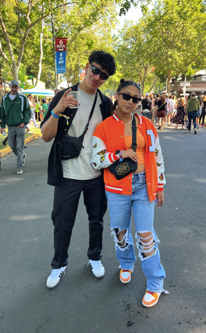 a man in a black and white outfit flanks a girl wearing an orange letterman jacket with butterflies on the sleeves with matching Air Force 1 sneakers