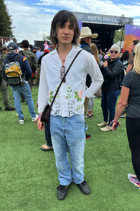 a male leaning individual wears a vintage white button down shirt with embroidered roses and lace detail plus jeans with studs scattered on the legs with a cross body bag and highlights in his hair