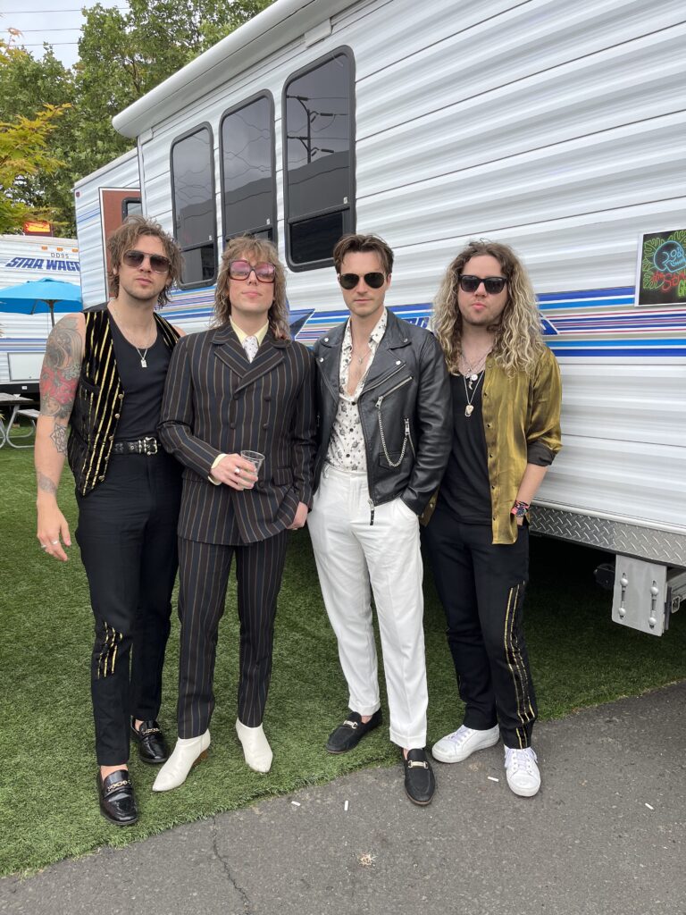 The 4 members of the band The Struts pose in stylish suits and garb in black and gold tones in front of their trailer