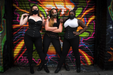 three femmes pose in front of a graffiti wall, wearing masks