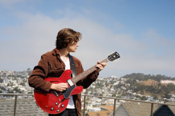 Musician Asher Belsky holds a Gibson guitar with the city of San Francisco behind him