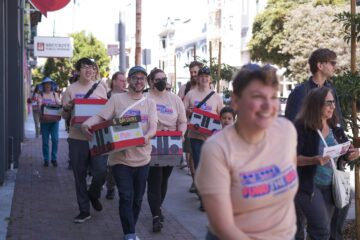 A group of smiling people carry boxes decorated like different Muni busses