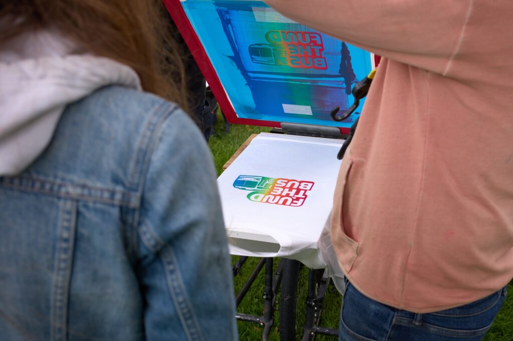 Someone is screenprinting a shirt that says "Fund the Bus" in rainbow colors 