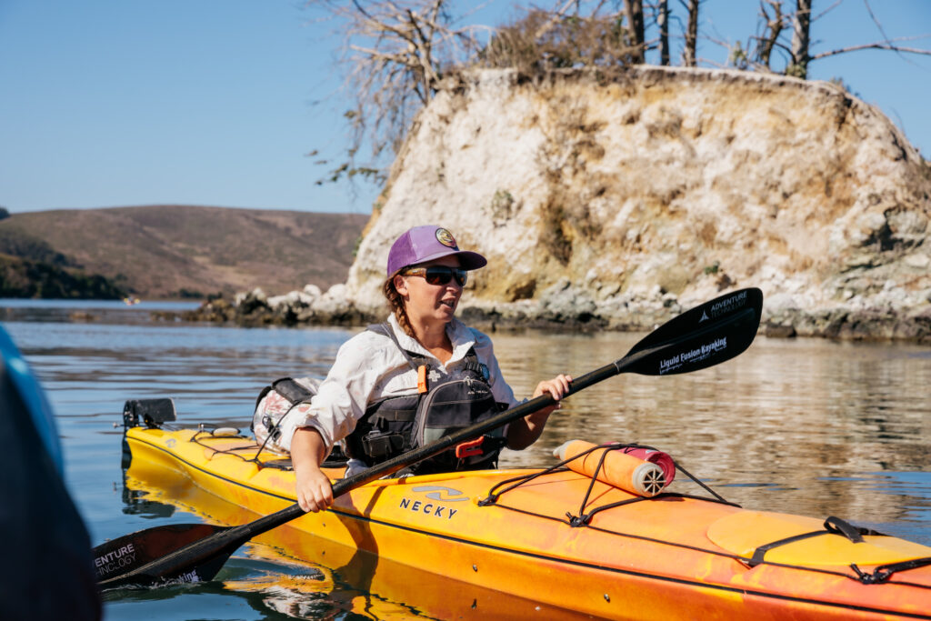 Photo of a woman wearing a purple hat in an orange kayak in front of an island. 